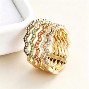 Può includere: Un anello color oro a più fasce composto da cinque file ondulate interconnesse. Ogni fila è incastonata con piccole pietre scintillanti di colori distinti, tra cui blu, verde, arancione, trasparente e viola, creando un effetto arcobaleno.