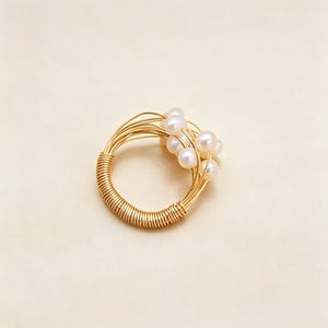 K&ouml;nnte beinhalten: Ein Ring aus goldfarbenem Draht mit einem Band aus eng gewickeltem Draht. Der obere Teil des Rings besteht aus mehreren d&uuml;nnen Drahtstr&auml;ngen, die sich auff&auml;chern und mehrere kleine, runde, wei&szlig;e Perlen in einer Cluster-Anordnung halten.