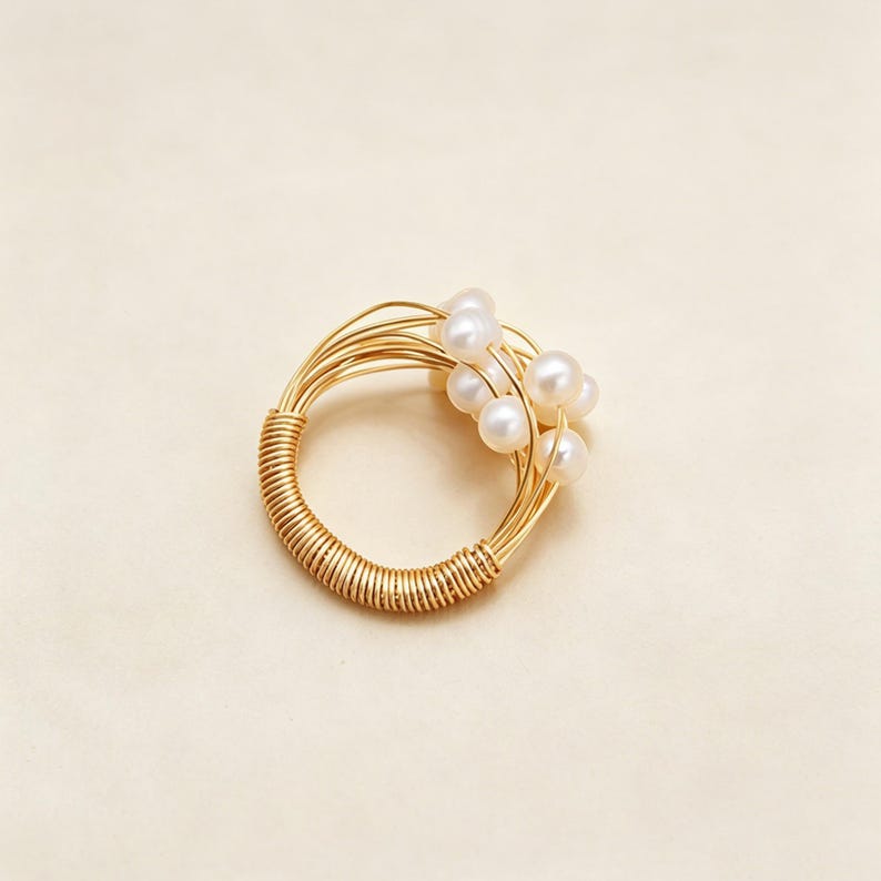 K&ouml;nnte beinhalten: Ein Ring aus goldfarbenem Draht mit einem Band aus eng gewickeltem Draht. Der obere Teil des Rings besteht aus mehreren d&uuml;nnen Drahtstr&auml;ngen, die sich auff&auml;chern und mehrere kleine, runde, wei&szlig;e Perlen in einer Cluster-Anordnung halten.