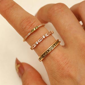 May include: Three gold-toned rings stacked on a finger. The top ring features small olive-green stones, the middle ring has alternating clear baguette and round stones, and the bottom ring is a wide band set with a row of olive-green square stones.
