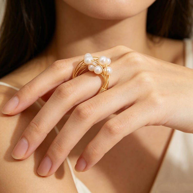 K&ouml;nnte beinhalten: Ein goldfarbener Ring aus mehreren d&uuml;nnen, gewickelten Metalldr&auml;hten, die sich um den Finger legen. Der Ring ist mit mehreren kleinen, runden, wei&szlig;en Perlen besetzt, die oben gruppiert sind und ein geschichtetes, strukturiertes Aussehen erzeugen.