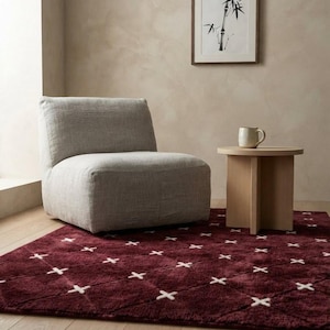 May include: A deep burgundy area rug featuring a white cross-stitch diamond pattern, paired with a light grey upholstered armless chair and a small round light wood side table holding a ceramic mug, set on a light-coloured wooden floor.