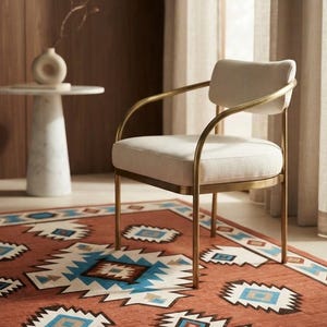 May include: An armchair with a curved backrest and seat upholstered in off-white fabric, supported by a thin, metallic gold-toned frame. The chair features slender armrests that connect to the backrest, creating a continuous, rounded silhouette.