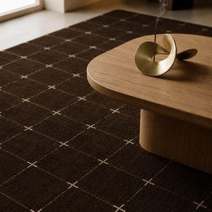 Puede incluir: Una alfombra de color marrón oscuro con un patrón de cuadrícula de líneas blancas finas con pequeñas cruces en cada intersección. Una mesa de centro de madera clara se asienta sobre la alfombra, sosteniendo un porta-incienso de metal dorado.
