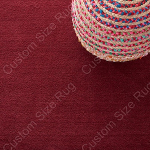 May include: A close-up of a deep red rug with a textured surface. A portion of a round, woven basket with colorful braided rope in shades of pink, blue, and green is in the upper right corner. The words "Custom Size Rug" are printed diagonally across the rug.
