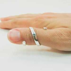 Könnte beinhalten: Ein offener Ring in polierter Silberoptik mit einer glatten, flachen Schiene, die sich teilweise um den Finger legt. Das Design umfasst zwei kleine, abgerundete, perlenartige Endstücke an beiden Seiten der Öffnung, die eine durchgehende Metallschleife bilden.