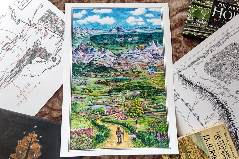 May include: A watercolor painting of a landscape with a winding road leading to a village in the distance. The scene is set in a valley surrounded by mountains and hills. The sky is blue with white clouds. The painting is framed in white.