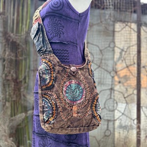 May include: A brown shoulder bag with embroidered circular designs and a button closure is displayed over a purple dress. The bag has a long strap and is made of a textured fabric. The dress has a sun and spiral design.