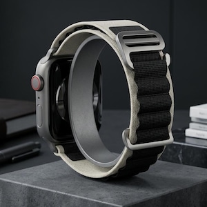 May include: A silver smartwatch with a black and off-white band is displayed on a gray stand. The watch has a red button and is next to a leather-bound notebook, a pen, and a stack of magazines.