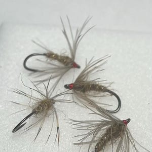 Op de afbeelding: Vier vliegvissen-kunstaasjes met gebogen metalen haken, beige synthetische lichamen met getextureerde vezels en dunne, uitstralende grijze hackle-veren. Elke vlieg is afgewerkt met een kleine, felrode kraal bij het oog van de haak.
