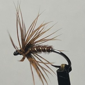 May include: A fishing fly lure featuring a dark brown body wrapped with thin gold wire. The lure has a cluster of long, thin, light brown and tan feathers extending outward from the front, attached to a curved black metal hook.