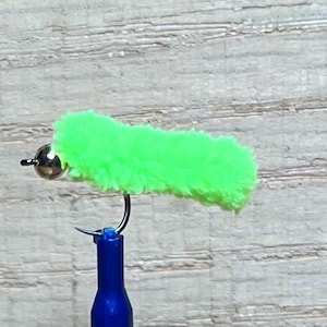 May include: A bright neon green fuzzy fishing fly lure featuring a metallic silver bead head attached to a curved metal hook. The lure is mounted on a blue cylindrical tool, showcasing the textured, soft material of the fly body clearly.