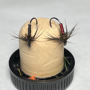 May include: Two fishing flies with dark hooks and thin, dark, wispy hackle fibres. One fly features a black ribbed body, while the other has a metallic red ribbed body. Both are mounted on a cylindrical tan cork plug set inside a black plastic cap.