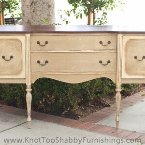 May include: A vintage-style, off-white wooden sideboard with a dark brown top. The sideboard has three drawers and two doors with ornate hardware. The sideboard is standing on a light brown floor.