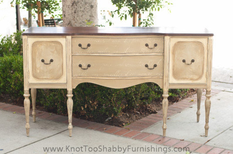May include: A vintage-style, off-white wooden sideboard with a dark brown top. The sideboard has three drawers and two doors with ornate hardware. The sideboard is standing on a light brown floor.