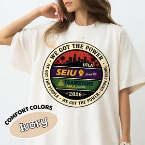 May include: Ivory-colored t-shirt with a circular graphic featuring text: "WE GOT THE POWER," "SEIU," "TEAMSTERS," and "2026." The graphic includes a cityscape silhouette and palm trees. The shirt is labeled "COMFORT COLORS."