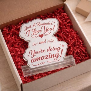 May include: A clear acrylic sign with the text "Just A Reminder: I Love You! and You're doing amazing!" in red script. The sign is in a white box filled with red paper shreds. A clear plastic stand holds the sign.