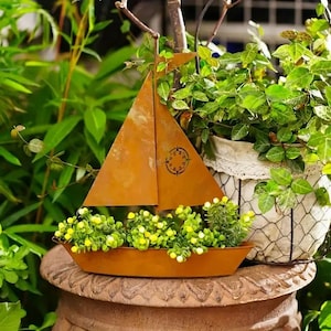 May include: A rusty brown metal sailboat planter filled with small green and yellow plants. The sailboat has a compass design on the sail and is surrounded by lush green foliage. The planter sits on a decorative stone pot.