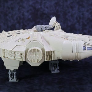 Może przedstawiać: Szczegółowy model statku kosmicznego Millennium Falcon, z jasnobeżową kolorystyką i skomplikowanymi panelami i detalami. Model zawiera antenę radarową, wieżyczki działek i podwozie, na ciemnoniebieskim tle.