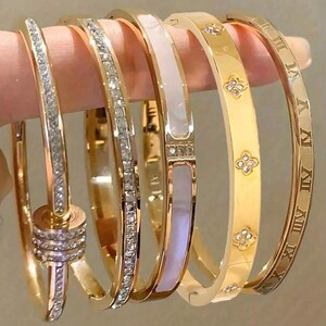 Könnte beinhalten: Ein Set aus fünf goldfarbenen Metallarmreifen. Einer verfügt über einen dreifachen Ringanhänger mit Kristallen, einer ist mit kleinen klaren Kristallen besetzt, einer hat eine weiße Einlage, einer zeigt vierblättrige Blumenmotive mit Kristallen.
