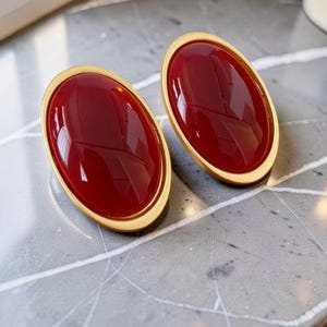 Könnte beinhalten: Ein Paar oval geformte Ohrringe mit glatten, tiefroten Cabochon-Mitten, die in polierte goldfarbene Metallrahmen gefasst sind. Die Ohrringe haben eine glänzende Oberfläche und ein schlichtes, abgerundetes Design, das den Kontrast zwischen Rot und Gold betont.