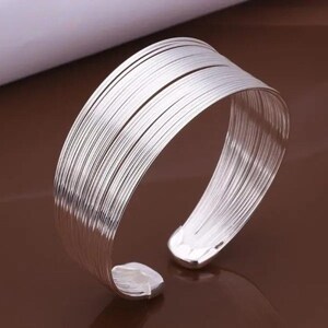 May include: A wide cuff bracelet composed of multiple thin, parallel silver-toned wire strands that wrap around the wrist. The bracelet features an open design with two rounded, solid end caps that secure the layered wire structure.