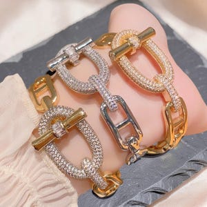 Könnte beinhalten: Drei Gliederarmbänder mit Knebelverschluss. Zwei Armbänder sind goldfarben mit klaren Pavé-Steinen besetzt, eines ist silberfarben mit klaren Pavé-Steinen. Jedes Armband besteht aus ovalen Gliedern mit einem zentralen Steg.