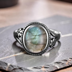 Könnte beinhalten: Ein silberfarbener Ring mit einem ovalen Labradorit-Edelstein in schillernden Blau- und Grautönen. Die Metallschiene ist auf beiden Seiten der zentralen Fassung mit kleinen Blumenmotiven verziert und hat eine texturierte, oxidierte Oberfläche.