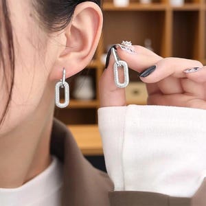 Könnte beinhalten: Ein Paar silberfarbene Ohrhänger, bestehend aus einem kleinen Klappcreolen-Verschluss, an dem ein längliches, ovales Glied befestigt ist. Das ovale Glied ist auf der gesamten Vorderseite mit kleinen, klaren, funkelnden Steinen besetzt.
