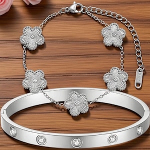 Könnte beinhalten: Ein silberfarbenes Schmuckset bestehend aus einem aufklappbaren Armreif und einem zarten Kettenarmband. Der Armreif ist mit klaren Steinen besetzt, während das Kettenarmband mit fünf silbernen Blumenanhängern mit texturierten Blütenblättern verziert ist.