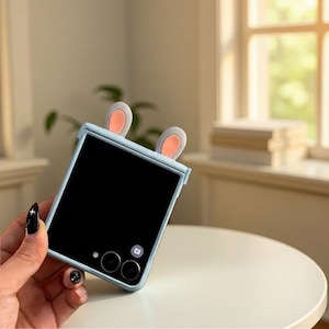 May include: A light blue phone case with attached bunny ears. The case has a black screen and a camera module. The bunny ears are light gray with pink inner ears. The phone case is held in a hand.
