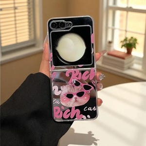 Puede incluir: Funda transparente para teléfono con un diseño rosa y negro. La funda presenta un gato de dibujos animados con gafas de sol, la palabra "Rich" y la palabra "Cat". La funda se muestra abierta, revelando la cámara y la pantalla del teléfono.