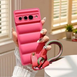 May include: A bright pink phone case with a quilted design. The case has a matching pink strap and a multi-colored wrist strap. The case has cutouts for the phone's camera and other features.