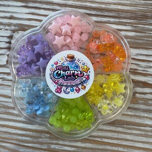 May include: A clear, flower-shaped container filled with colorful charms. The charms include stars, beads, and flower shapes in shades of pink, orange, yellow, green, blue, and purple. The container has a circular label with the text "Mini Charm Lab."