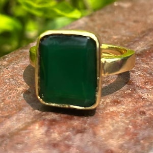 Op de afbeelding: Een goudkleurige ring met een grote, rechthoekige, smaragdgroene edelsteen. De edelsteen is gezet in een gouden bezel en de ringband is ook goudkleurig. De ring wordt getoond tegen een getextureerde, bruine ondergrond met groen gebladerte op de achtergrond.