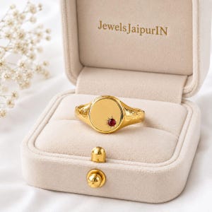 Può includere: Un anello sigillo dorato con fascia martellata e superficie tonda piatta che presenta una piccola pietra rossa incastonata a stella. L'anello è esposto all'interno di un portagioie in velluto beige con il testo JewelsJaipurIN stampato in oro.