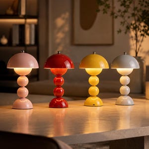 May include: Four table lamps in pink, red, yellow, and grey, each with a mushroom-shaped design. The lamps have a spherical base and a rounded top. The lamps are on a light-coloured surface.