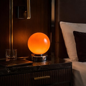 May include: An orange globe lamp with a chrome base sits on a dark marble nightstand. A glass of liquid and a small leather wallet are also on the nightstand. The lamp is next to a bed with white and brown pillows.