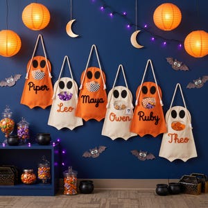 May include: Six hanging ghost-shaped treat bags in orange and cream felt, each featuring a cutout mouth for candy storage and a personalized name in felt lettering. The bags are displayed against a dark blue wall decorated with paper bats, crescent moons, and glowing orange paper lanterns.