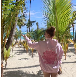 Könnte beinhalten: Hellrosa Kapuzenpullover mit einem Muscheldesign auf dem Rücken. Das Bild zeigt den Hoodie, der an einem Sandstrand getragen wird, mit Palmen und blauem Himmel im Hintergrund. Das Muscheldesign ist in einem dunkleren Rosa gehalten.
