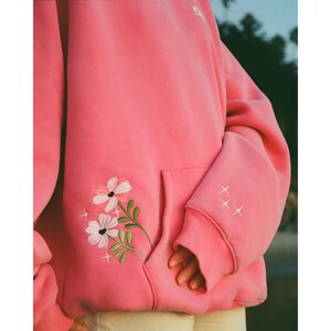 May include: A pink hoodie with a kangaroo pocket and ribbed cuffs. The hoodie features an embroidered floral design on the front and three embroidered stars on the sleeve. The garment is made of a soft material.