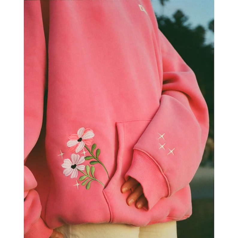 May include: A pink hoodie with a kangaroo pocket and ribbed cuffs. The hoodie features an embroidered floral design on the front and three embroidered stars on the sleeve. The garment is made of a soft material.