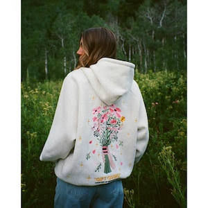 May include: A pale grey hoodie featuring a floral bouquet on the back. The bouquet includes pink, yellow, and white flowers tied with a green ribbon. The text "GIFT GIVING" is below the design.