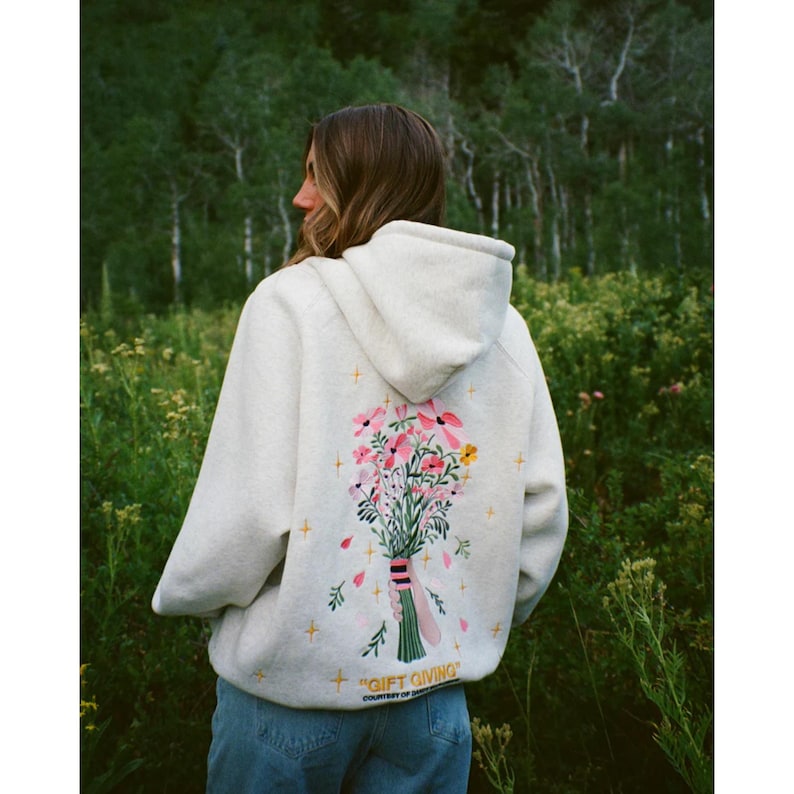 May include: A pale grey hoodie featuring a floral bouquet on the back. The bouquet includes pink, yellow, and white flowers tied with a green ribbon. The text "GIFT GIVING" is below the design.