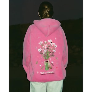 May include: A bright pink hoodie with a floral bouquet embroidered on the back. The bouquet features white and pink flowers with green stems and leaves. The text "GIFT GIVING" is below the bouquet.
