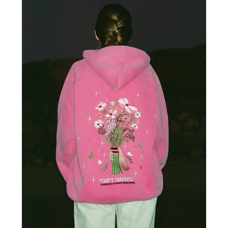 May include: A bright pink hoodie with a floral bouquet embroidered on the back. The bouquet features white and pink flowers with green stems and leaves. The text "GIFT GIVING" is below the bouquet.