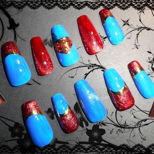 Pode incluir: Um conjunto de dez unhas postiças com um esquema de cores azul e vermelho vibrante com detalhes em glitter. Algumas unhas incluem um emblema dourado estilo Superman ou uma faixa dourada horizontal. O design é inspirado num super-herói.