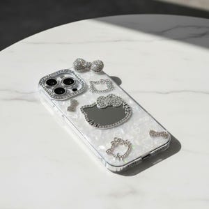 May include: A white phone case with a pearlescent finish, decorated with sparkling rhinestones. The case has a silver frame around the camera and rhinestone bows and Hello Kitty designs.