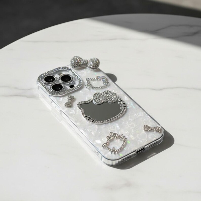 May include: A white phone case with a pearlescent finish, decorated with sparkling rhinestones. The case has a silver frame around the camera and rhinestone bows and Hello Kitty designs.