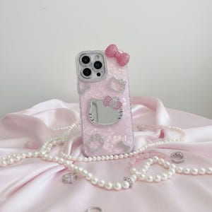 May include: A pink phone case featuring a Hello Kitty design, adorned with rhinestones and a bow. The case includes a mirror and is surrounded by pearl necklaces and rings, displayed on a pink satin fabric.